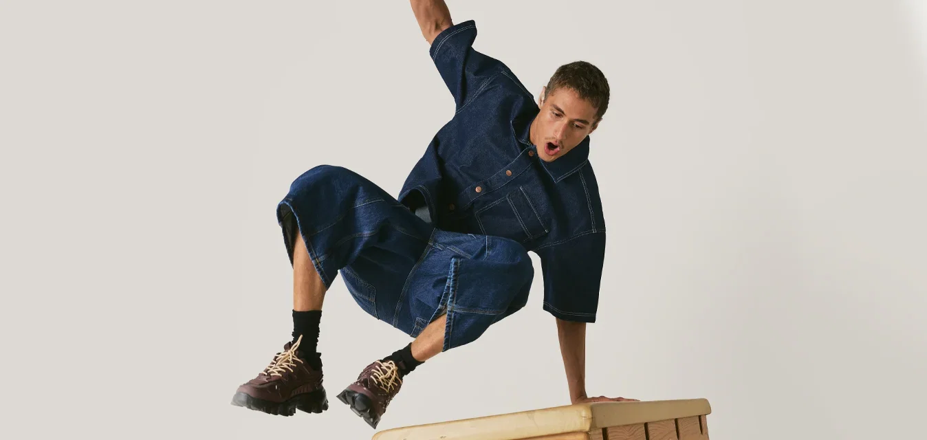 Man in spijkeroutfit springt over een houten gymnastiekbok met één hand die zijn val ondersteunt.