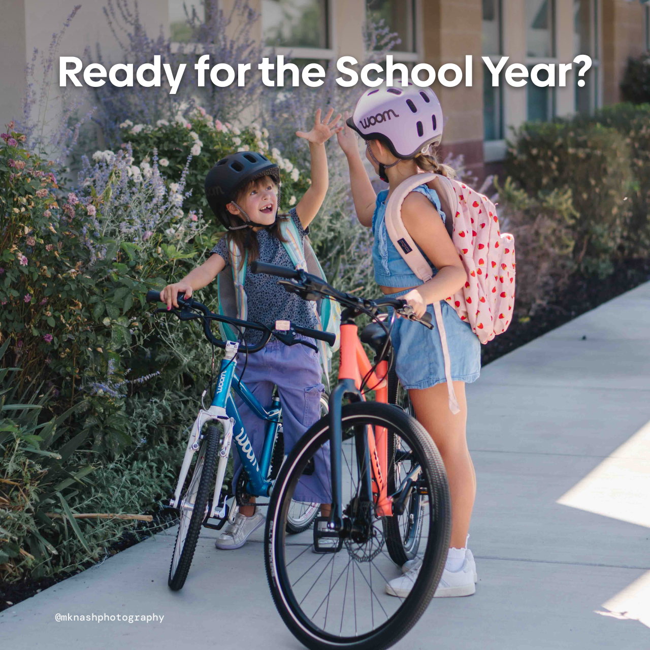Text over photo reads "Ready for the School Year?" with image showing two girls high fiving while standing with their woom GO and EXPLORE bikes in front of their school. Both wear backpacks and READY Kids' Helmets.
