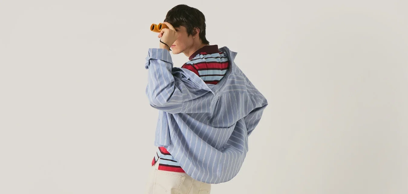 Persona con camicia a righe chiare e pantaloni beige che guarda attraverso un binocolo arancione su sfondo uniforme.