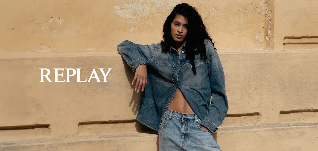 Femme aux longs cheveux bouclés appuyée contre un mur beige texturé, portant une veste en jean, un jean large et des baskets beiges à l'extérieur.
