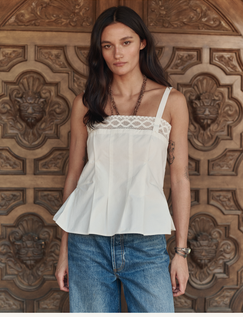 A woman with long dark hair wears a white sleeveless top with lace detail and blue jeans, standing in front of an ornate wooden door.