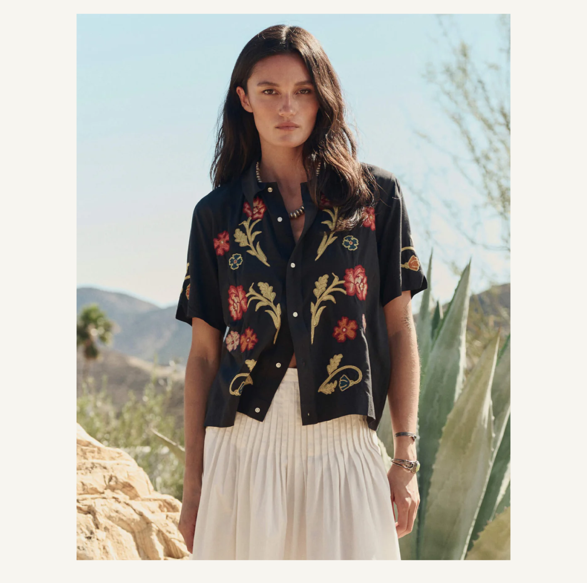 A woman with long dark hair wears a black shirt with colorful floral embroidery and a white skirt, standing outdoors near agave plants and rocks with mountains in the background.