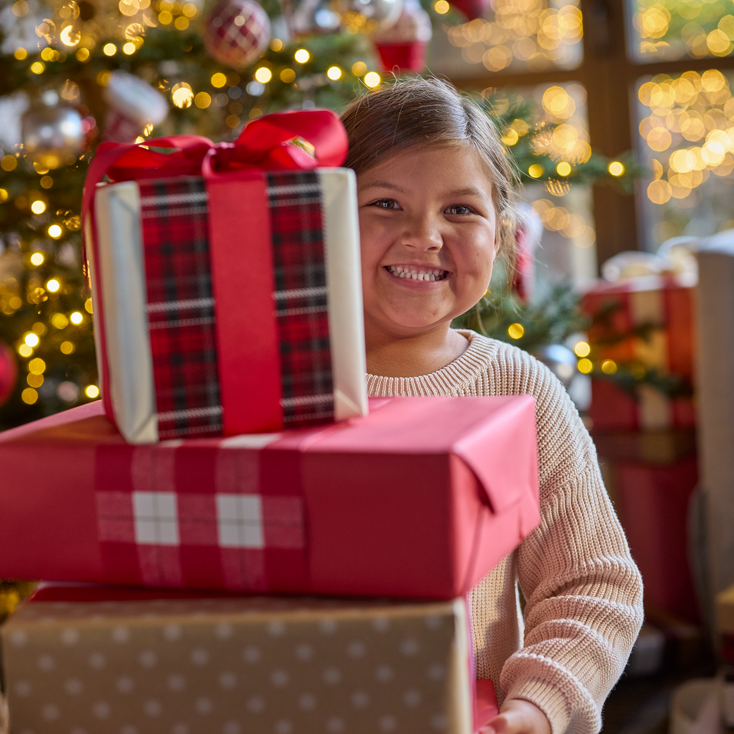Smiling child standing behind wrapped Christmas gifts with a decorated tree and warm lights in the background.