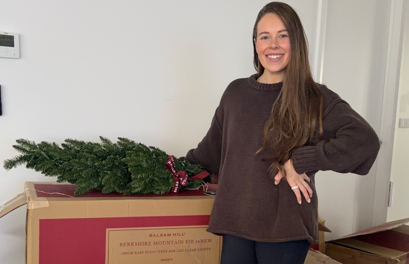 Woman standing next to a boxed artificial Christmas tree with greenery placed on top.