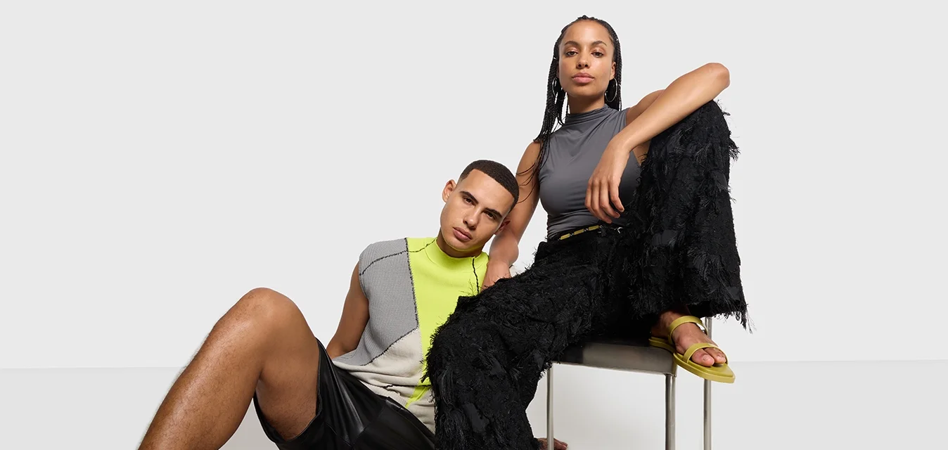 Dos modelos con ropa moderna posan contra un fondo liso; uno está sentado en una silla con pantalones negros y sandalias amarillas, mientras que el otro se reclina a su lado.