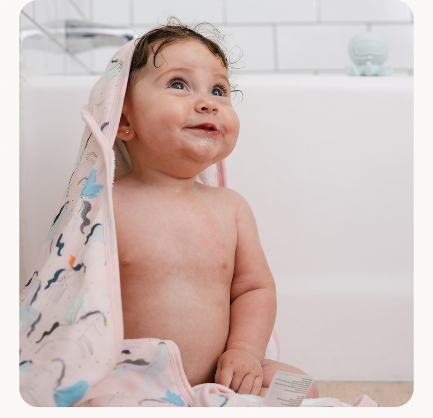 baby girl with dark hair and pierced ears sitting near a bathtub