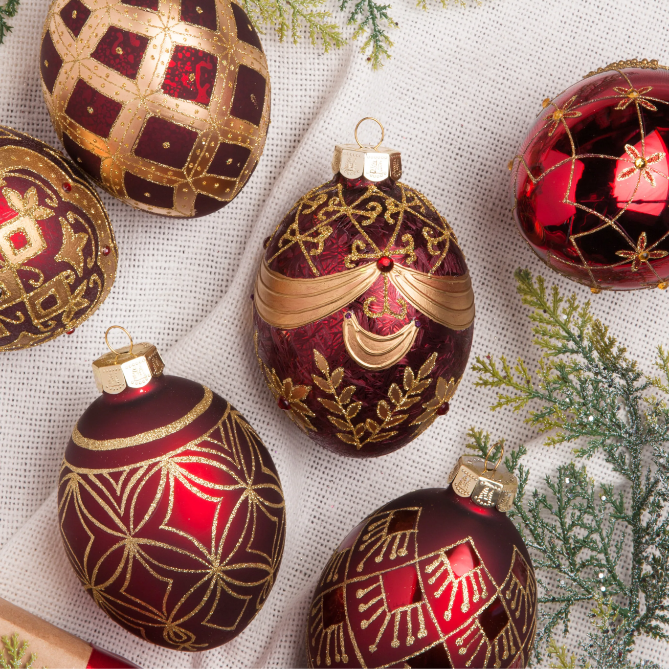 Close-up of red and gold Christmas ornaments with intricate patterns on a white fabric background.
