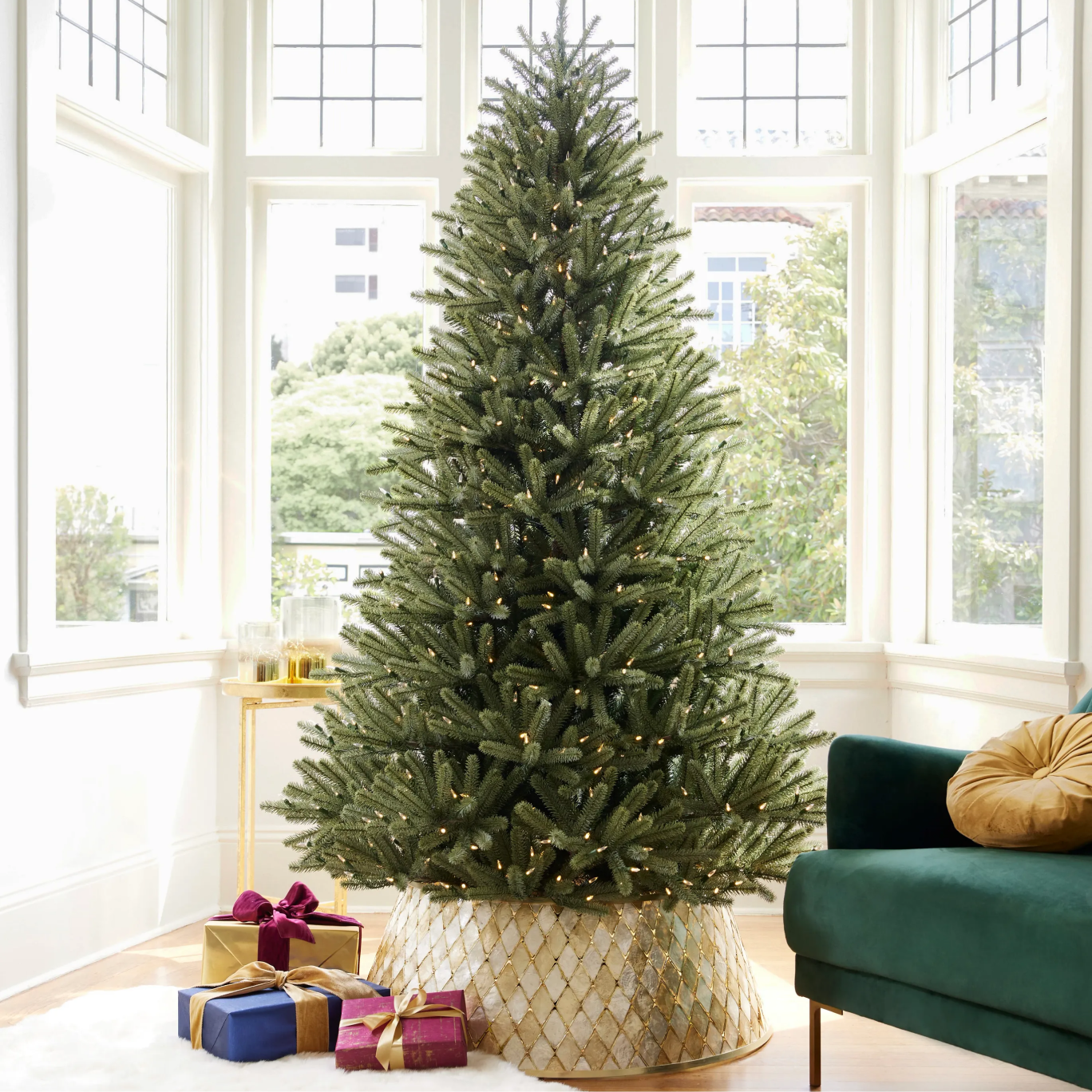 Pre-lit artificial Christmas tree displayed in a bright room with large windows, wrapped gifts, and a gold-patterned tree collar.
