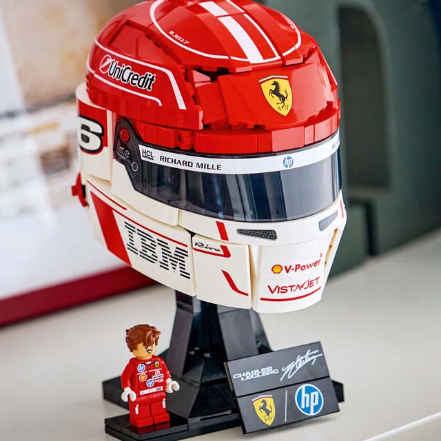 Red and white LEGO helmet with logos, mounted on a stand, accompanied by a small LEGO figure in a racing suit.
