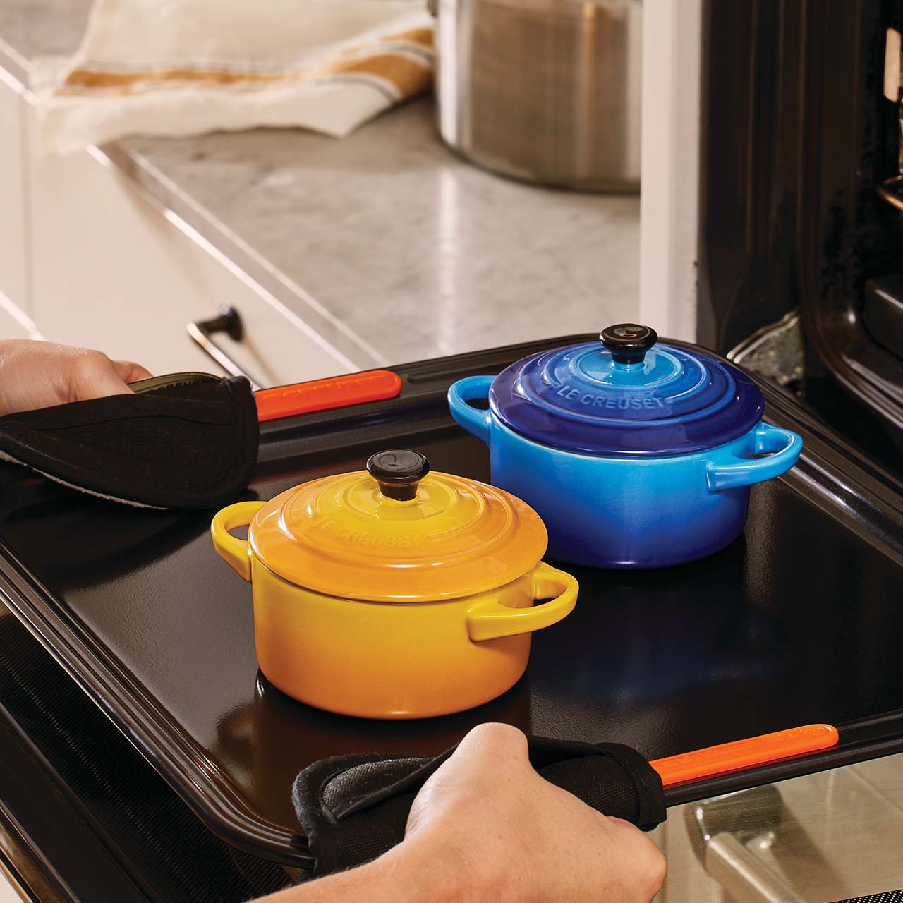 Two small colorful pots, one yellow and one blue, are placed on a black oven tray being removed from an oven.