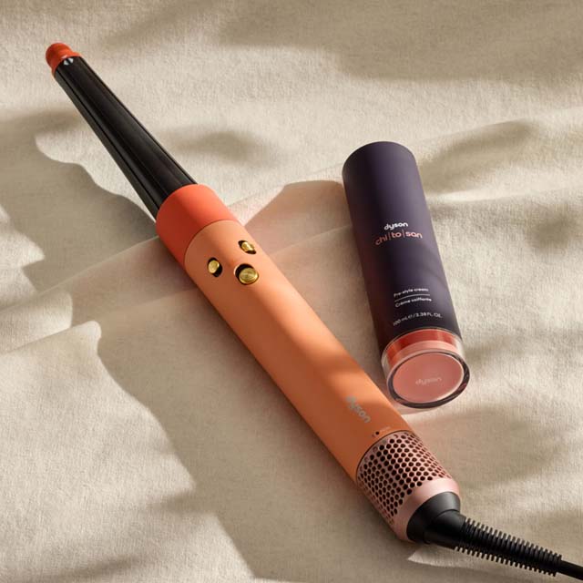 A curling iron and a purple cylindrical container are placed on a light fabric surface.