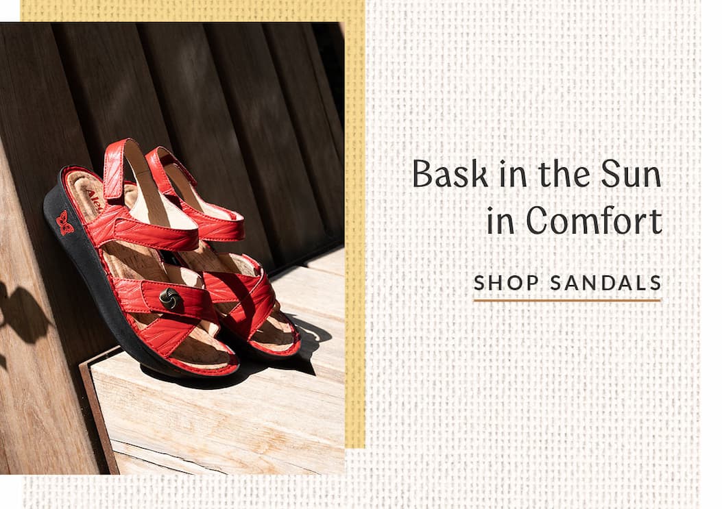 A pair of vibrant red strappy sandals with black soles are resting on light-colored wooden steps under bright sunlight. On the right side, text on a light textured background reads "Bask in the Sun in Comfort" above an underlined "SHOP SANDALS" link.
