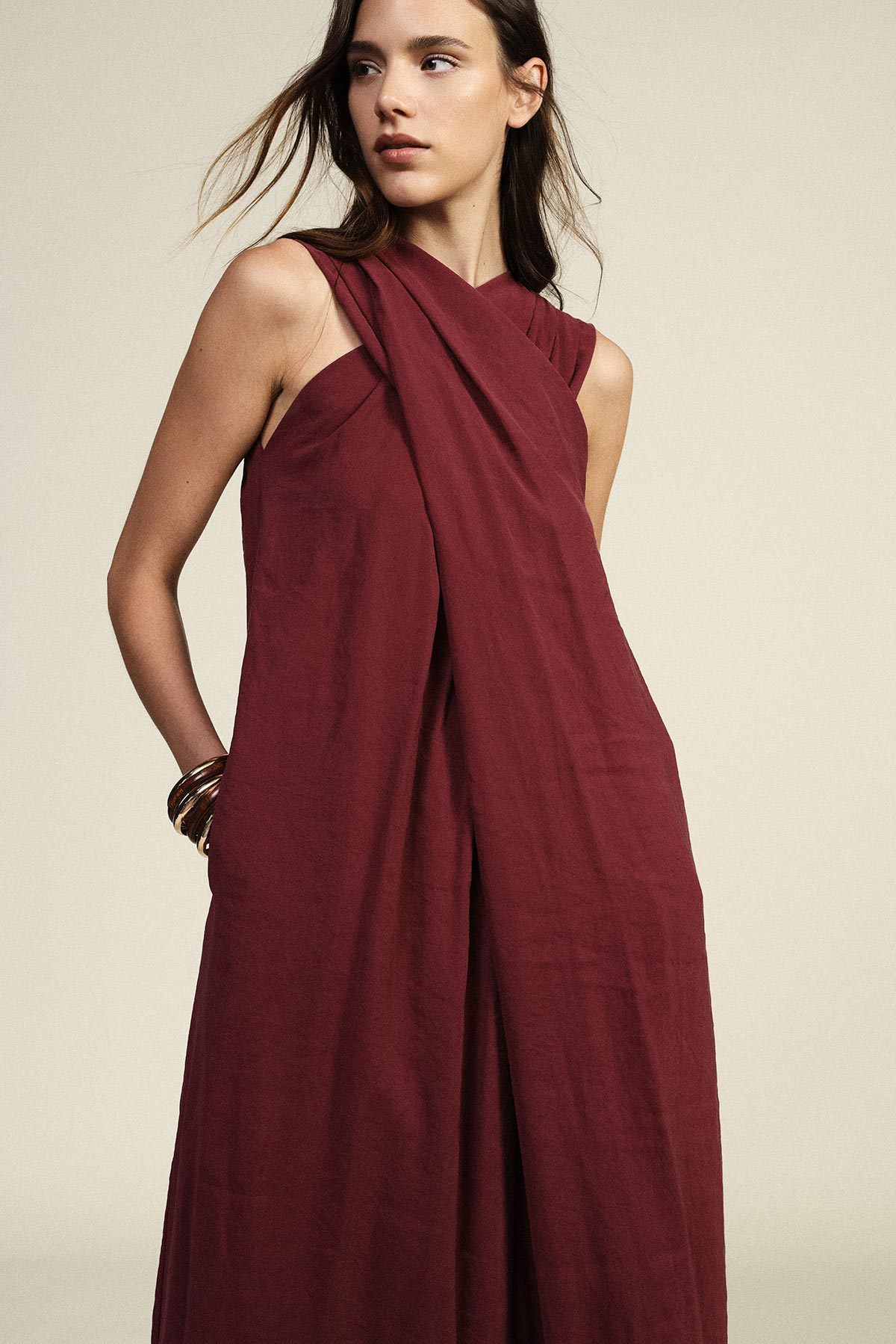 Woman wearing sleeveless burgundy draped maxi dress with criss-cross neckline, hands in pockets and gold bangles