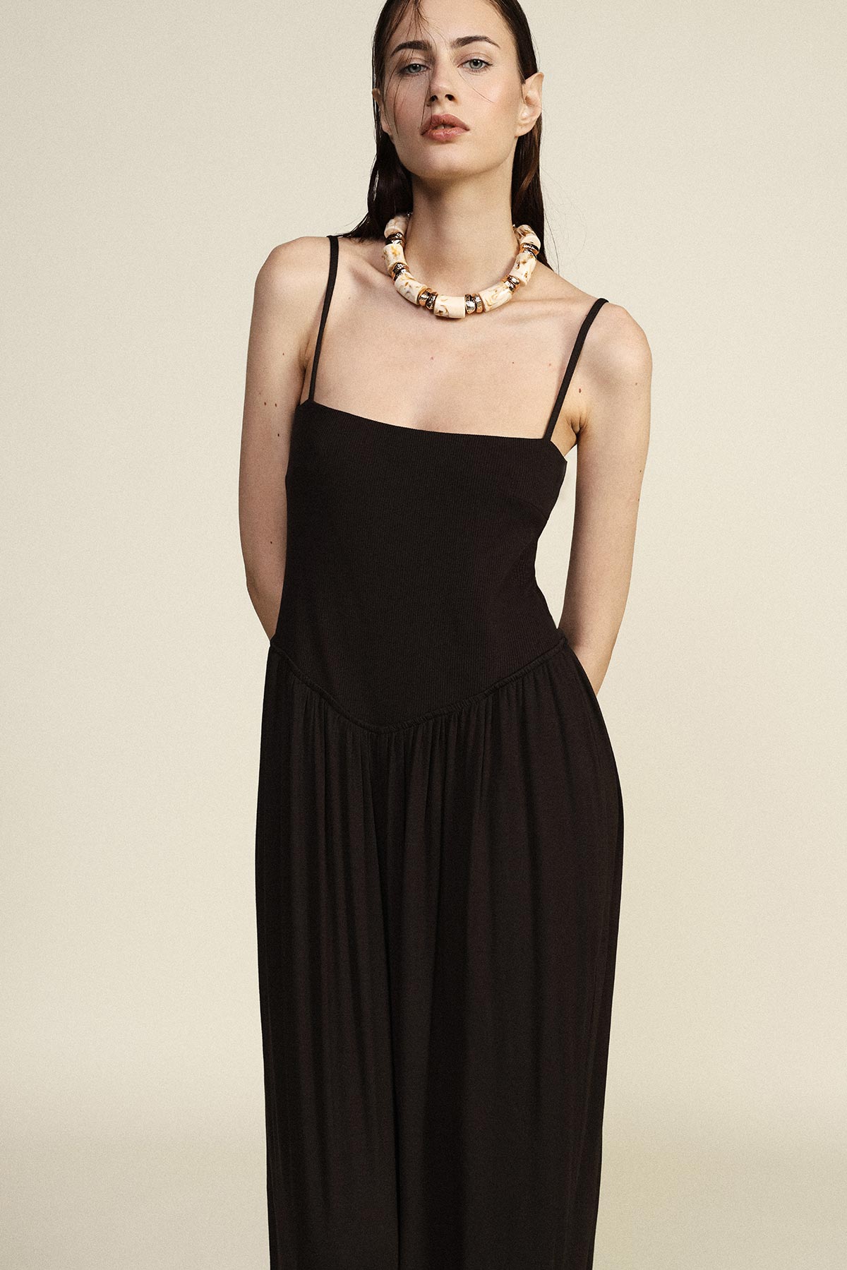 Woman wearing a black spaghetti-strap maxi dress and chunky beige statement necklace against a neutral background