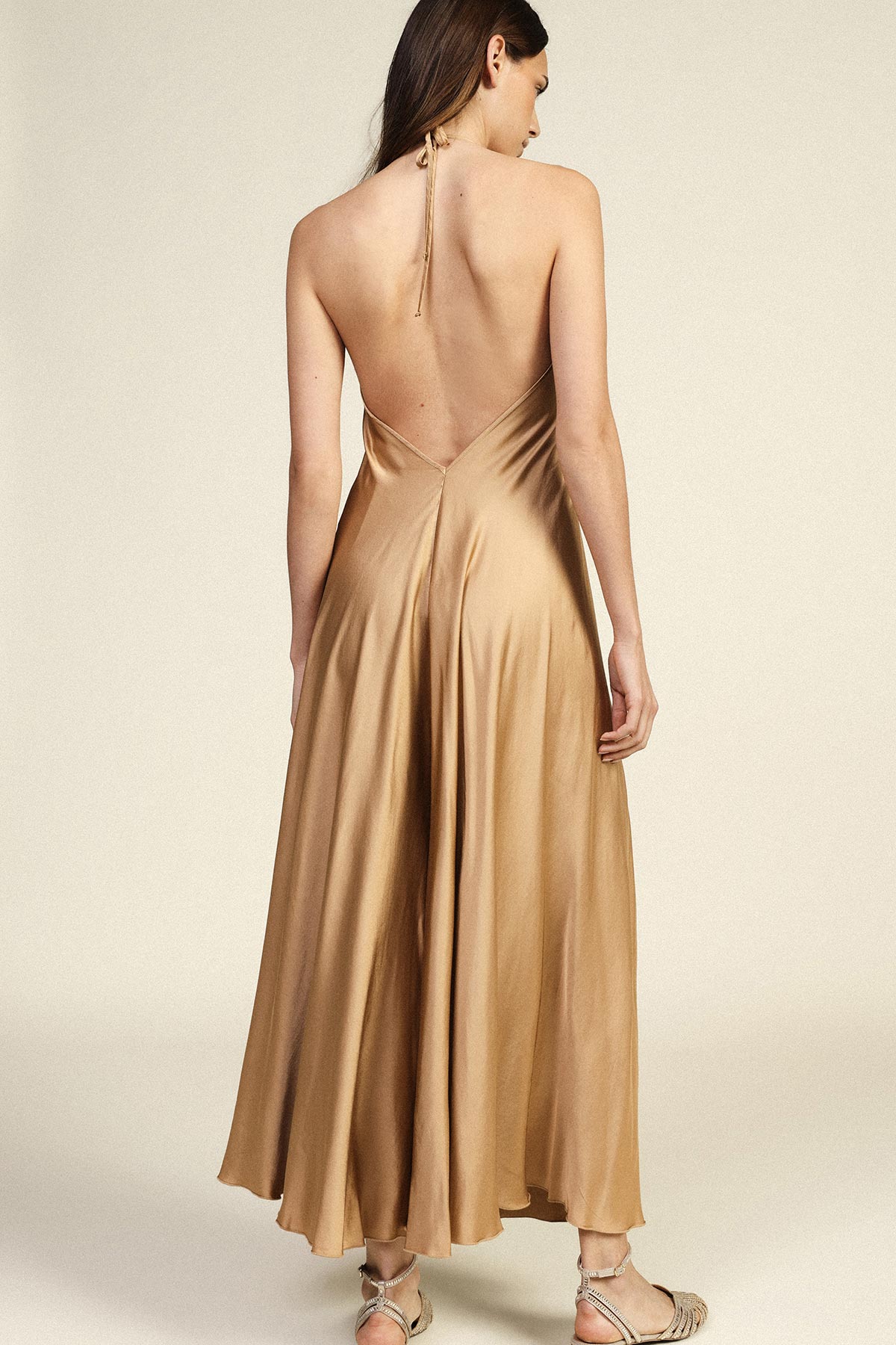 Back view of woman wearing a gold satin backless halter dress with flowing skirt and strappy sandals