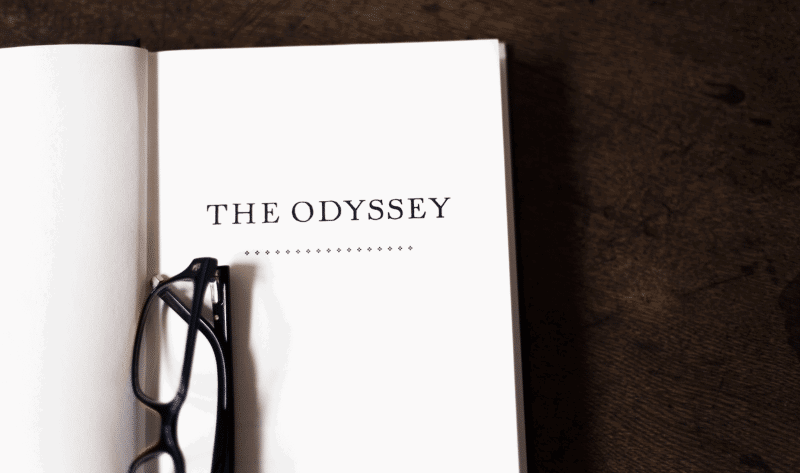Open book displaying the title "The Odyssey" on a white page, with a pair of black eyeglasses resting on the left side, set on a dark wooden surface.