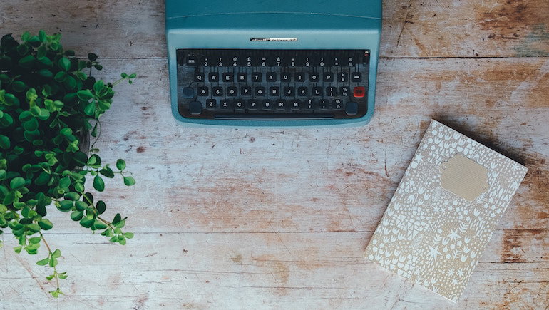 A blue typewriter sits on a wooden table beside a potted plant and a beige patterned notebook.