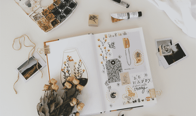 Open scrapbook with dried flowers, assorted drawings, Polaroid photos, color palette, paint tube, and other artistic supplies scattered on a white surface.