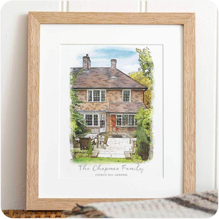 A framed watercolour illustration of a red brick house personalised with the family name, displayed in a wooden frame on a surface