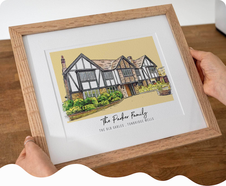 A framed watercolour illustration of a stone cottage personalised with the family name, held up by someone in a linen shirt beside a window with a plant in the background