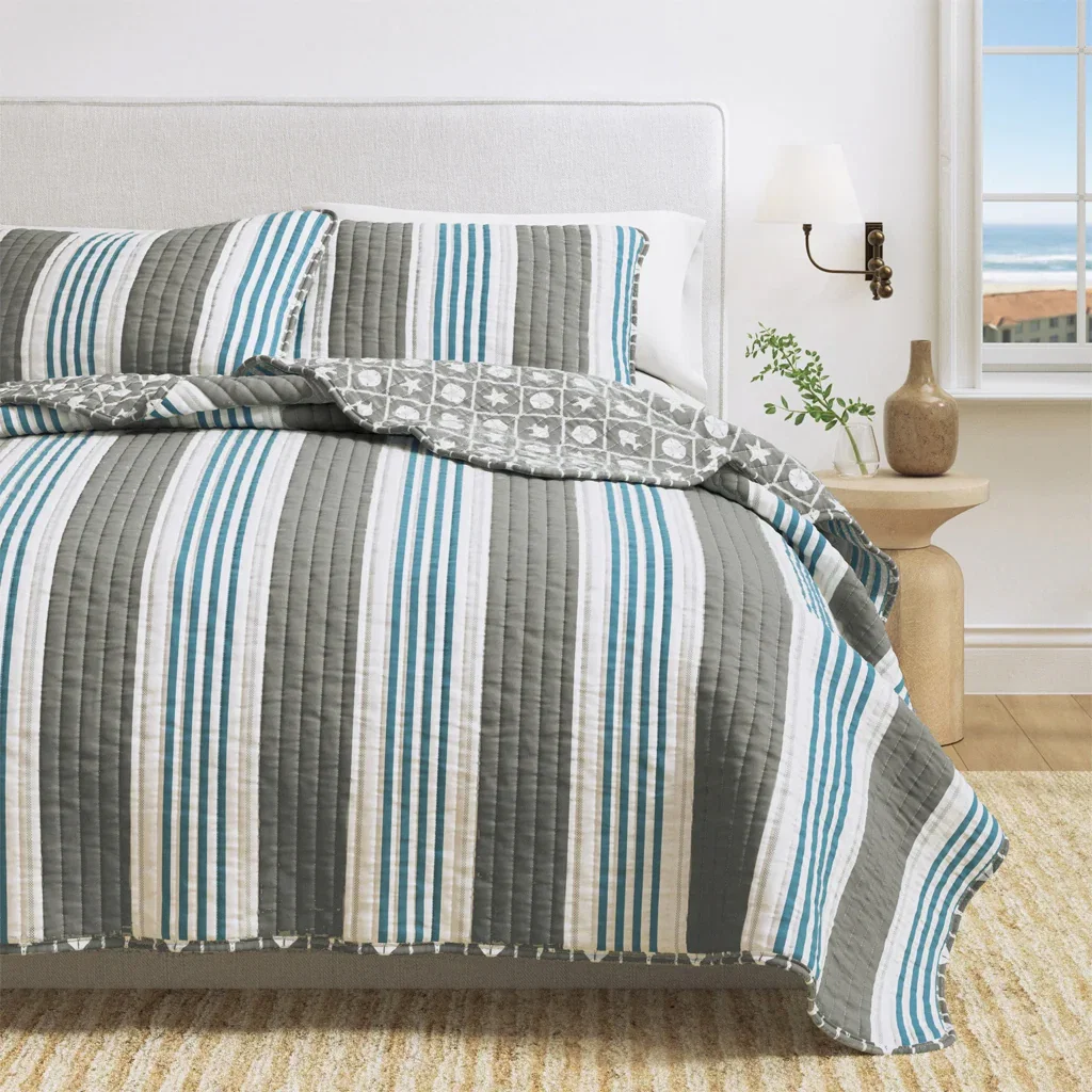 Striped quilt set in gray, white, and teal on bed with coastal decor and ocean view window.