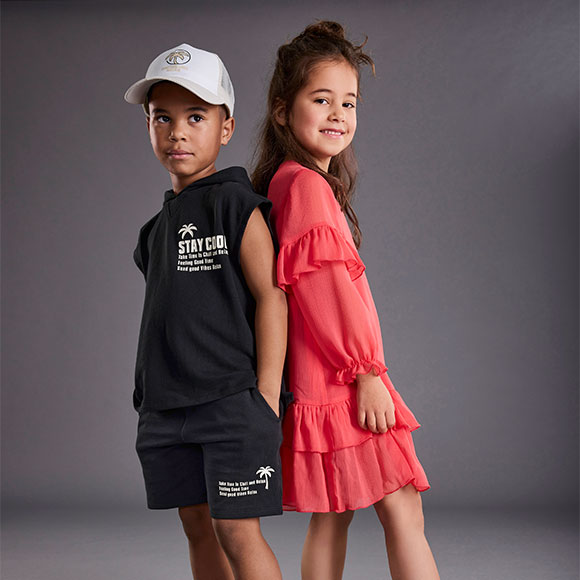 Junge trägt schwarzes T-Shirt mit weißem Aufdruck und schwarze Shorts, Mädchen trägt rotes Kleid mit Rüschen, Vorderansicht