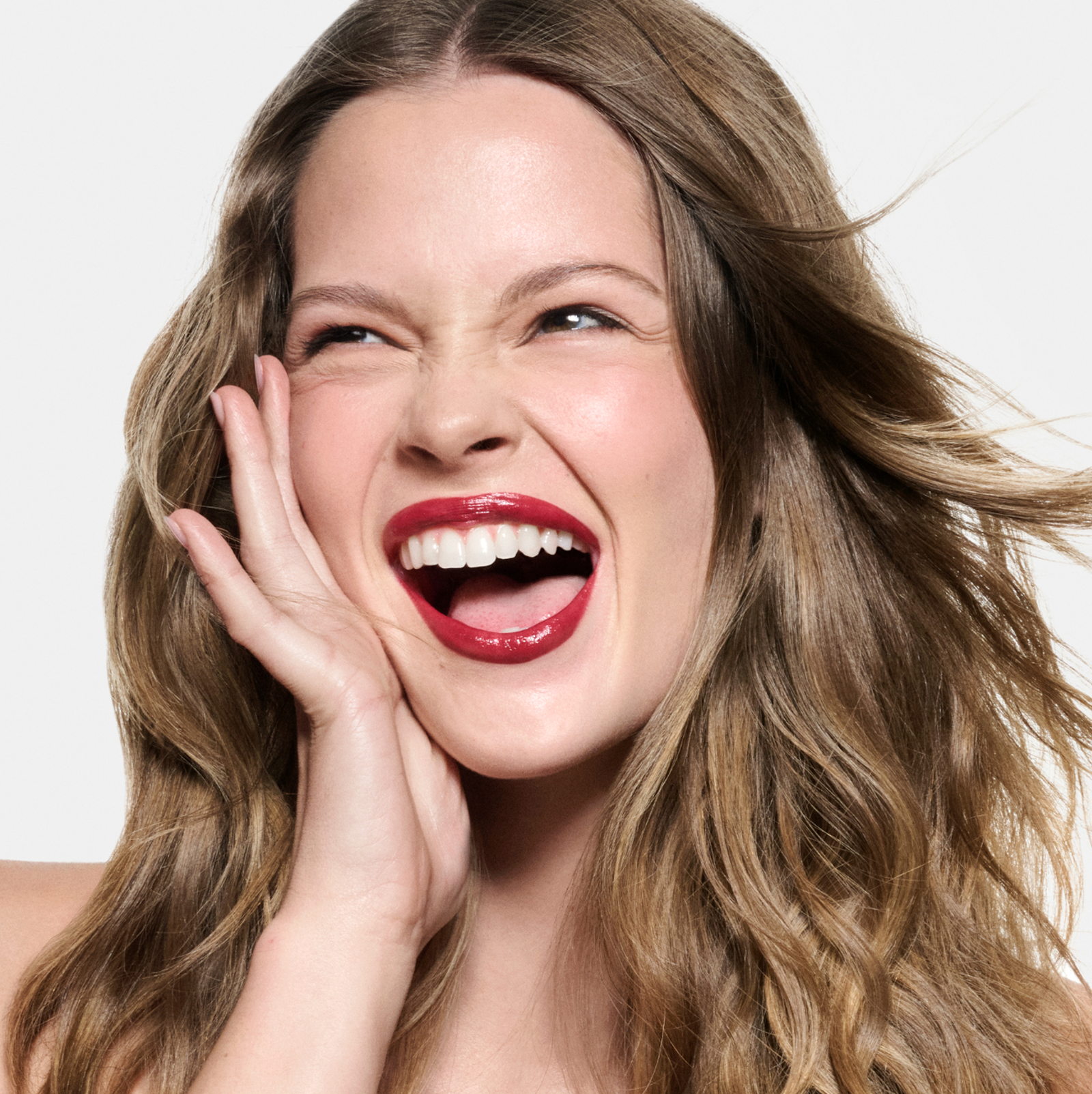 Close-up of a smiling woman with long, wavy brown hair wearing Haus Labs Ruby Atomic Shake Lip Lacquer, holding her hand to her cheek against a white background.