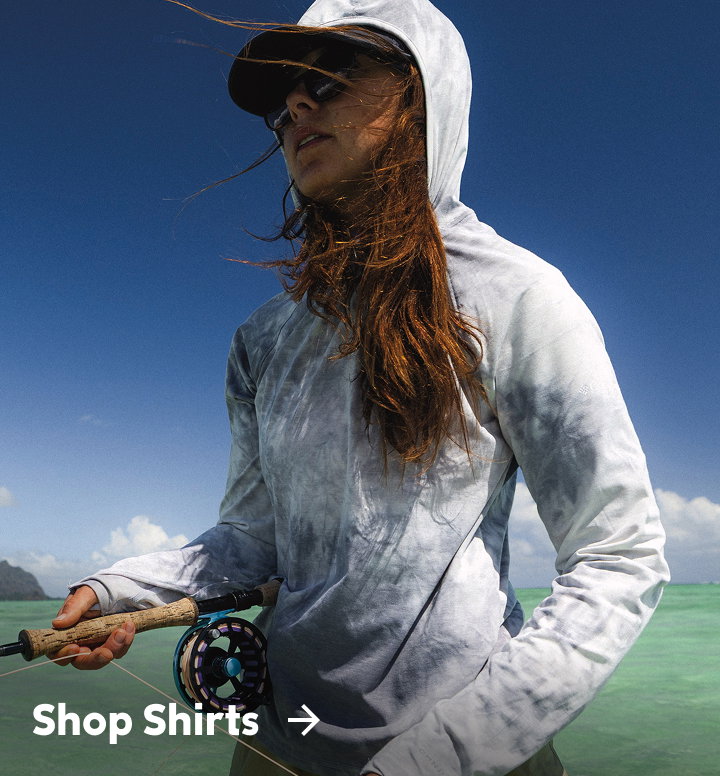 A woman standing in the water wearing a blue sun hoodie and holding a fishing rod with text: Shop Shirts
