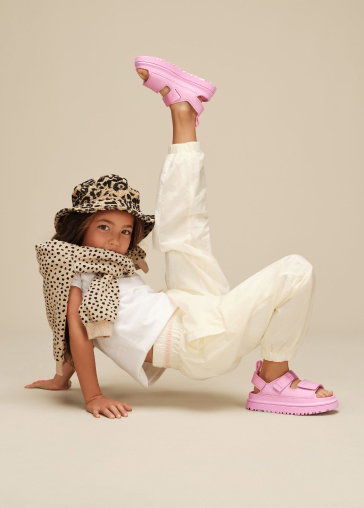 A studio shot of a model posing with a leg in the air, wearing a hat, t-shirt, trousers and kids' GoldenGlow Sandals