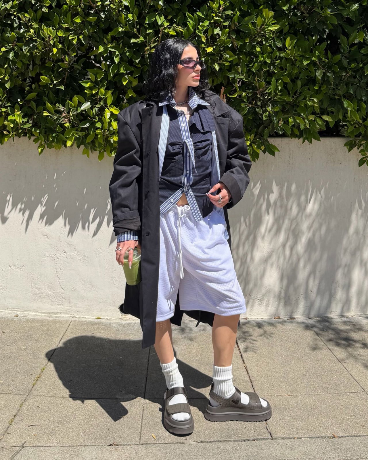 Person in black coat, light shirt, babyblue shorts, white socks with the goldenglow embossed sandal, sunglasses, holding a matcha.