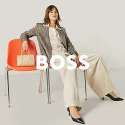 Femme en tenue professionnelle assise de manière détendue sur deux fauteuils orange, tenant un sac à main beige sur un fauteuil, sur un fond clair et uni.