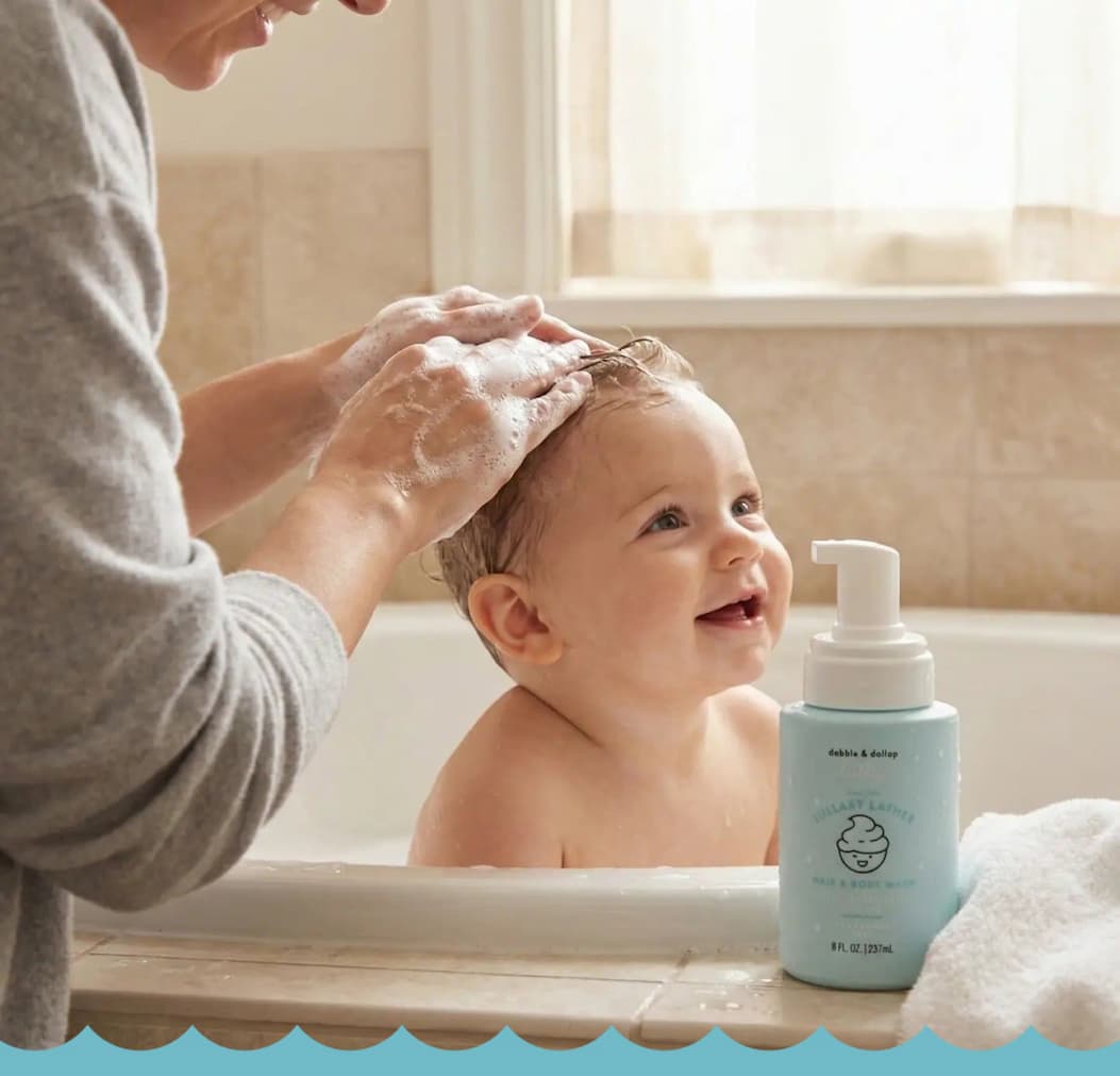 A joyful baby in a tub is having hair washed with foamy soap.