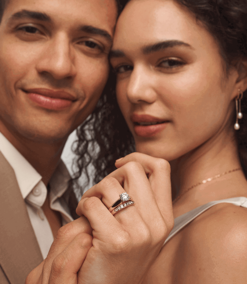 Close-up of woman wearing yellow gold pearl drop earrings and diamond bridal rings, next to a male model.