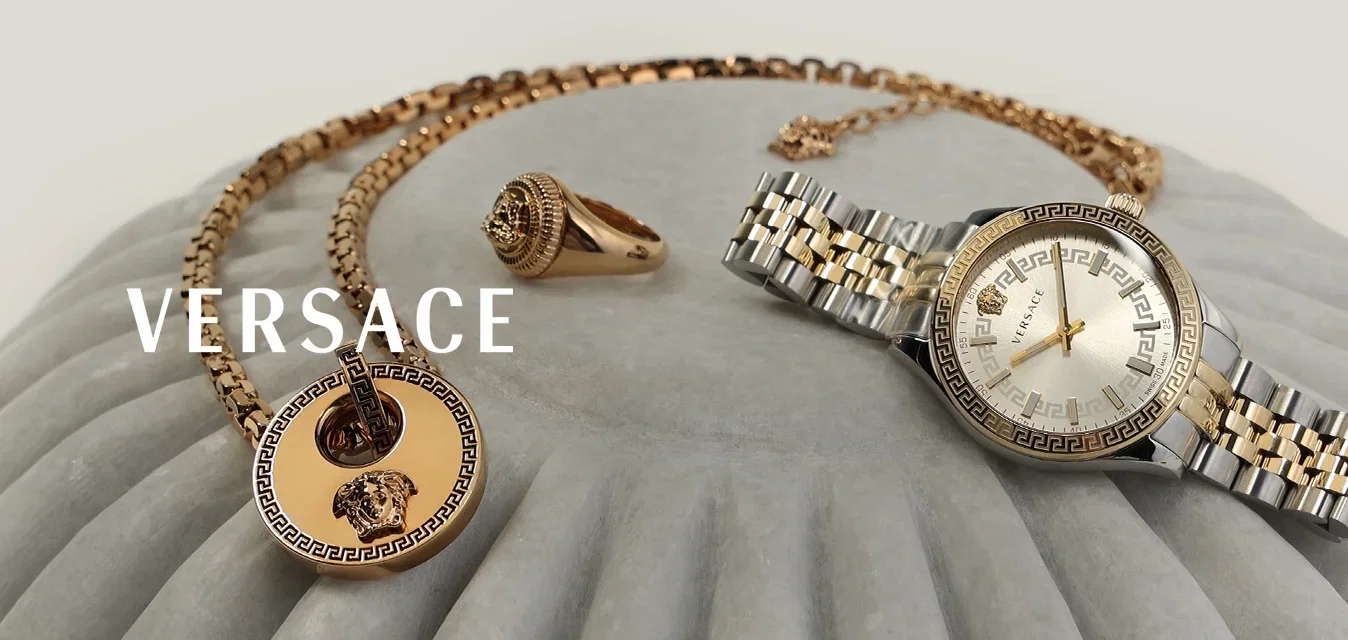 Collana d'oro con ciondolo circolare, anello d'oro con pietra e orologio da polso in metallo bicolore esposti su una superficie grigia scanalata.