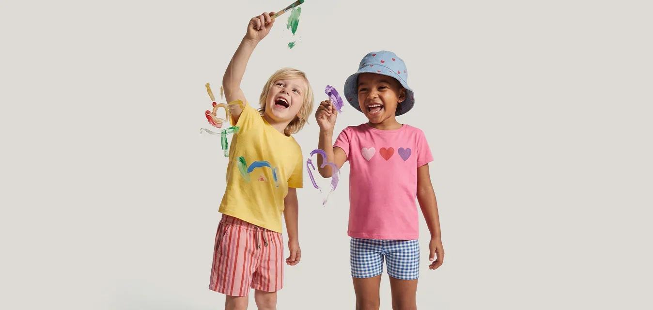 Dos niños pequeños sonriendo y sosteniendo molinillos de colores, uno lleva una camiseta amarilla y pantalones cortos rosas, el otro una camiseta rosa, sombrero azul y pantalones cortos.