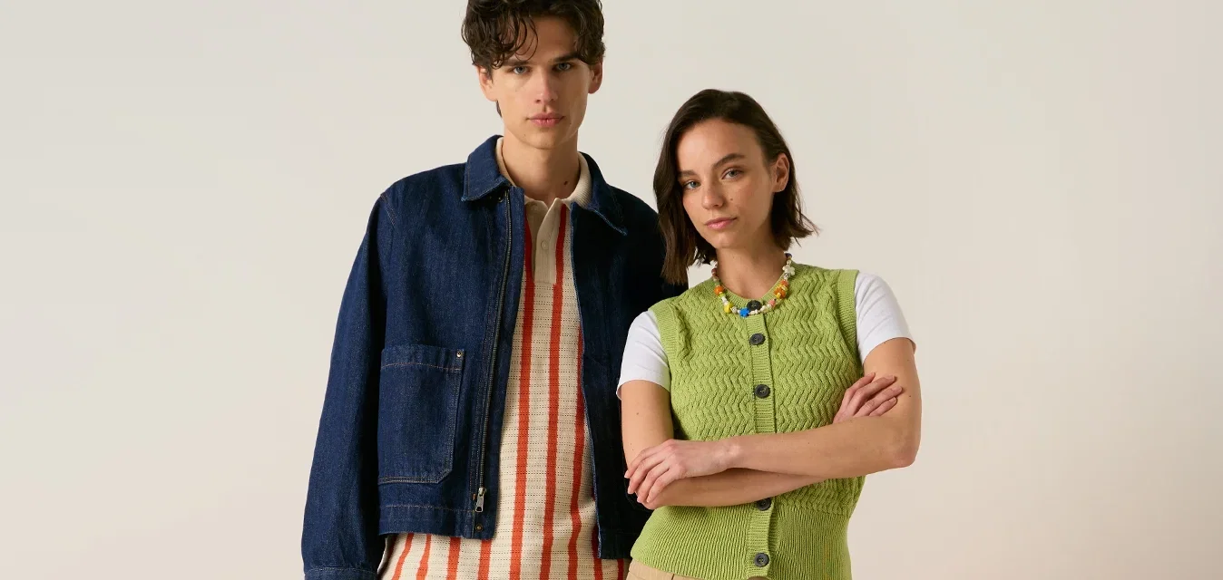 Joven hombre y mujer de pie frente a sillas naranjas, vestidos con ropa casual de primavera y zapatillas, fondo neutro.