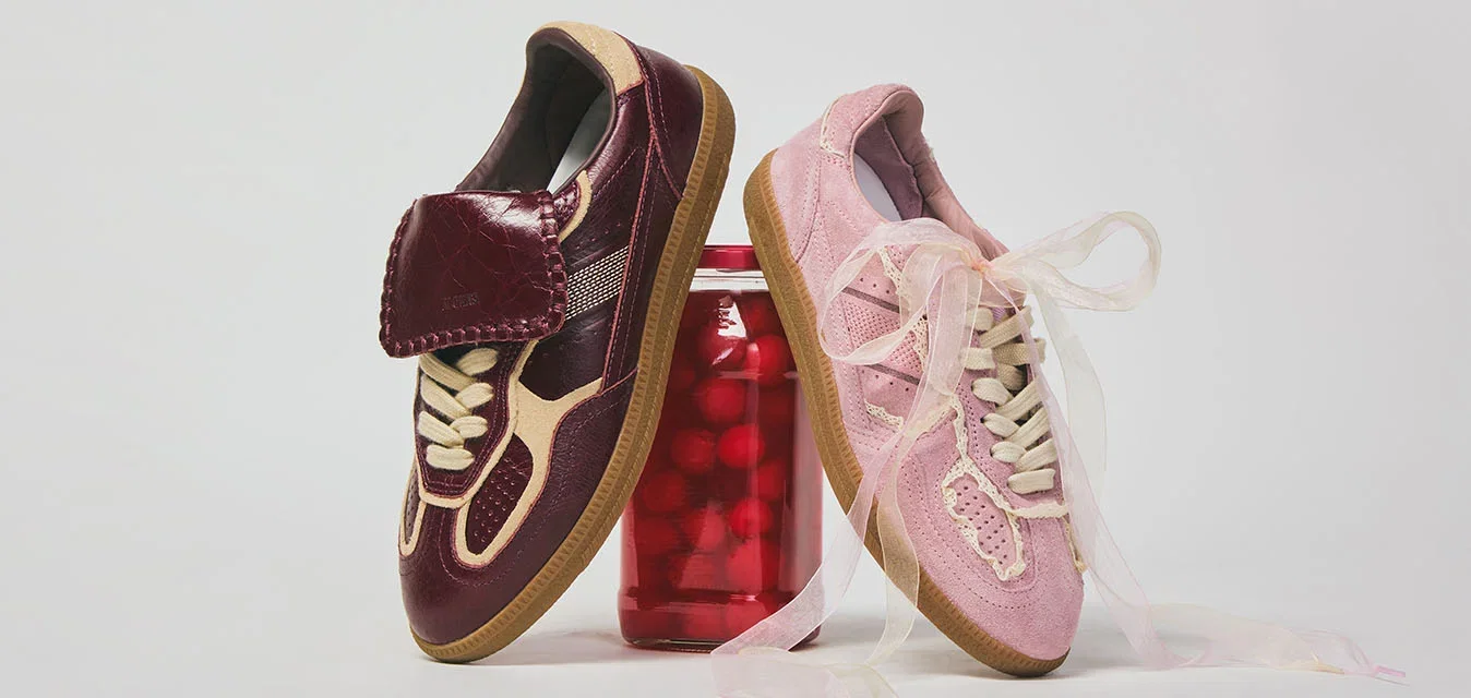 Dos zapatillas de estilo vintage, una rojo oscuro con una correa y otra rosa con cordones, equilibradas sobre un tarro rojo contra un fondo blanco liso.