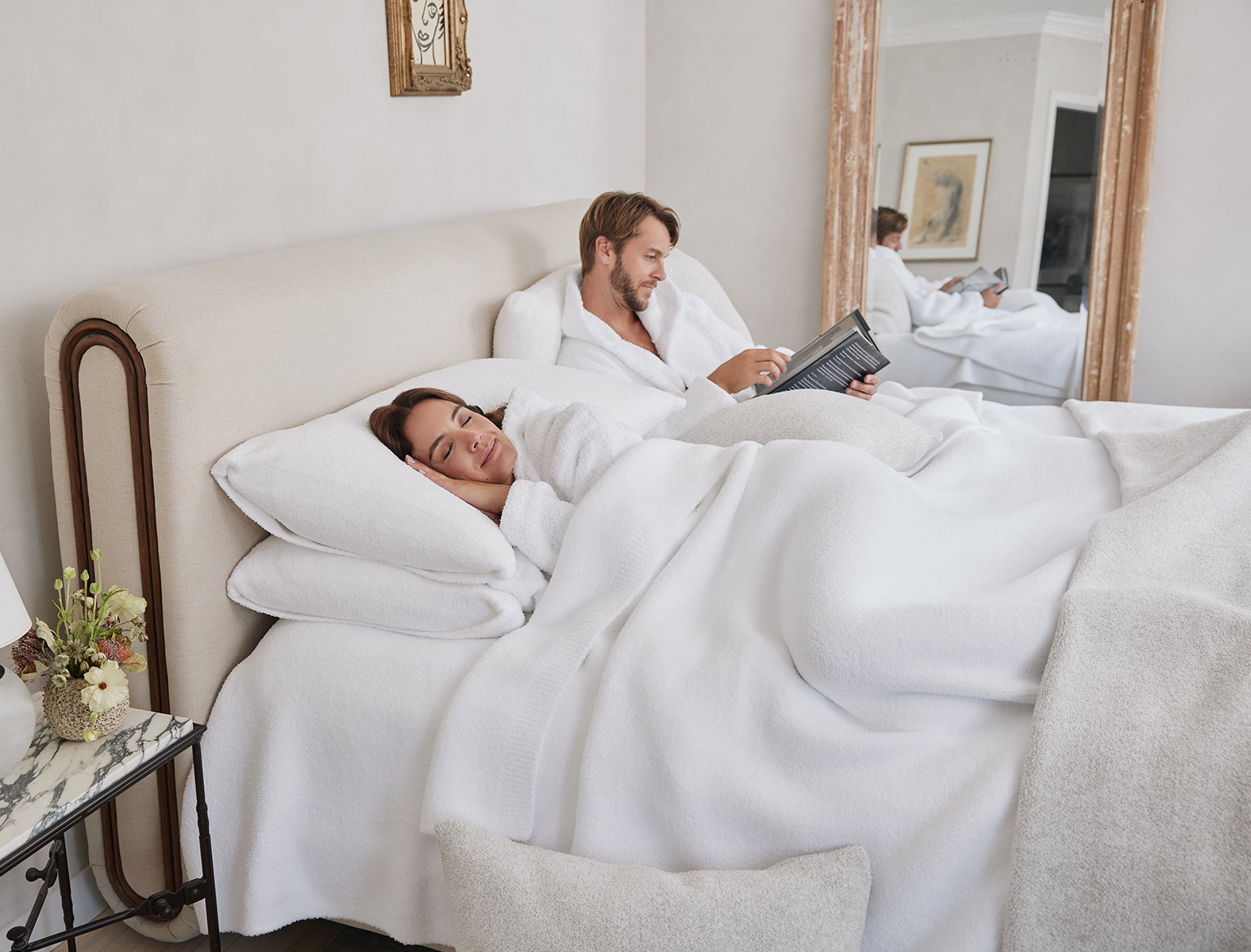 A couple relaxes in bed wrapped in plush white Kashwére blankets and robes, with layered pillows creating the ultimate cozy Sunday morning retreat.