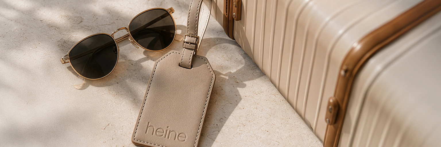  Eine Werbeanzeige mit dem Text Auf dem Bild ist kein Text zu sehen. Der Hintergrund zeigt eine Sonnenbrille, einen beigen Koffer mit goldfarbenen Akzenten und einen beigen Kofferanhänger mit der Prägung 'heine' auf hellem Untergrund.