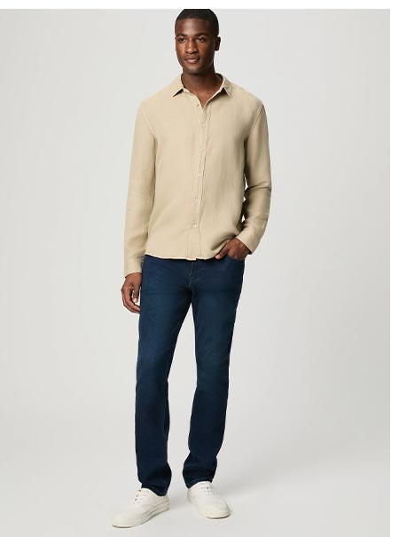 A man wearing a beige button up shirt and dark denim jeans.