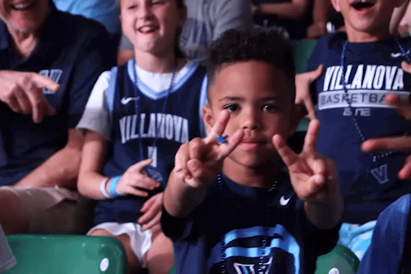 A young boy wearing a basketball jersey smiling and making peace signs in the stadium crowd.