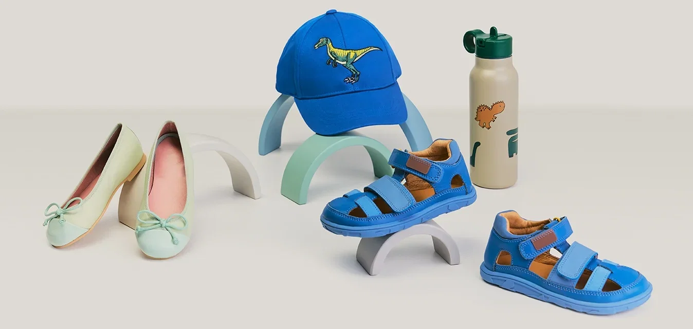 Par de bailarinas verdes pálidas para mujer, sandalias azules para niños, gorra azul con dinosaurio y botella de agua beige con dibujos de dinosaurios sobre superficie blanca.