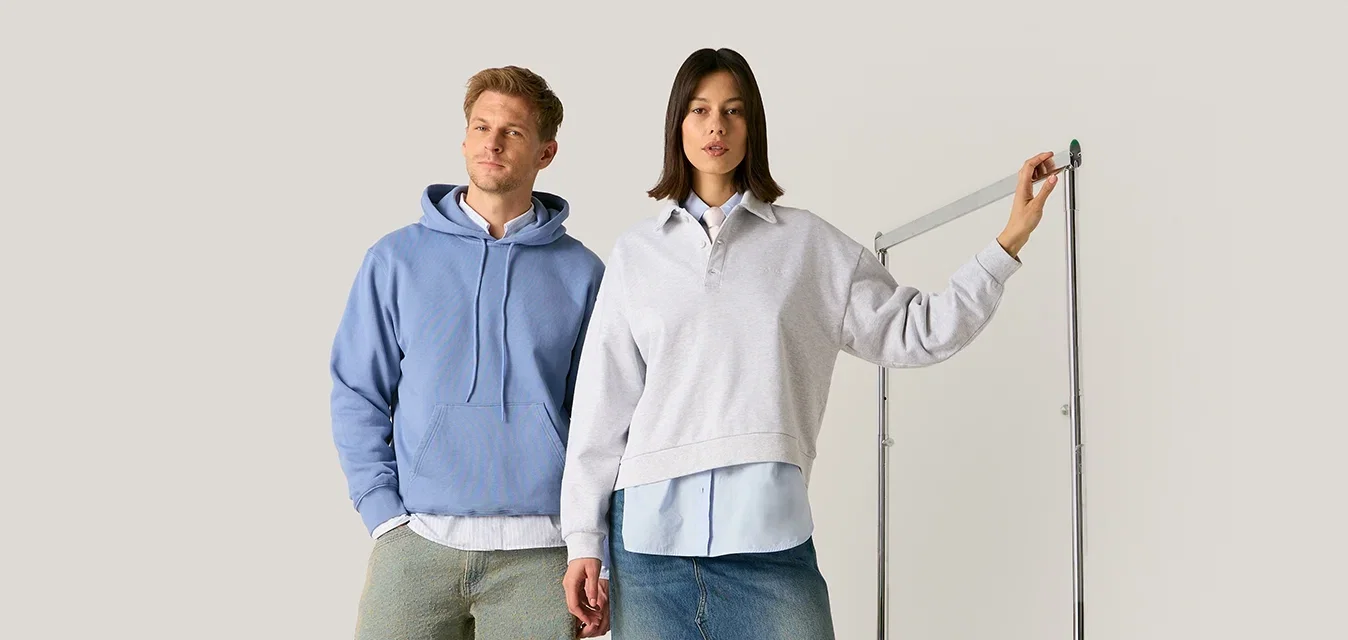 Hombre con sudadera azul con capucha y pantalones verdes y mujer con sudadera blanca y falda vaquera de pie, mujer sosteniendo dispositivo para medir la altura.