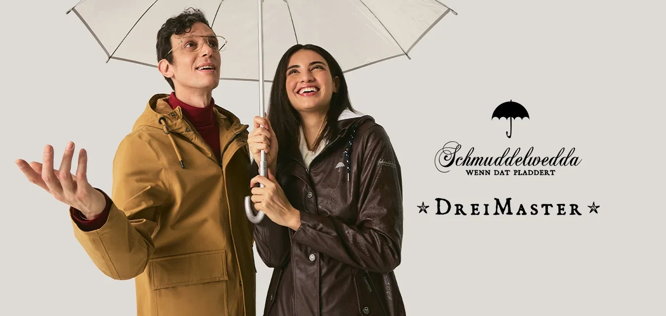 Hombre y mujer sonriendo bajo un paraguas transparente, vestidos con impermeables, comprobando si llueve contra un fondo claro y liso.