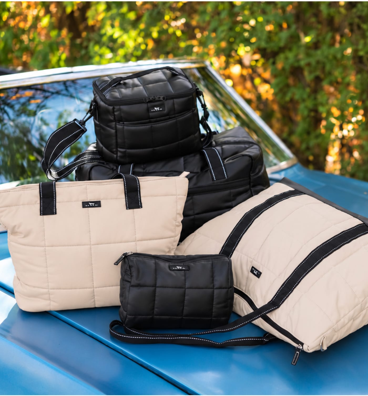 SCOUT. SCOUT. SCOUT. SCOUT. SCOUT. Collection of black and beige quilted SCOUT bags arranged on the hood of a blue car.