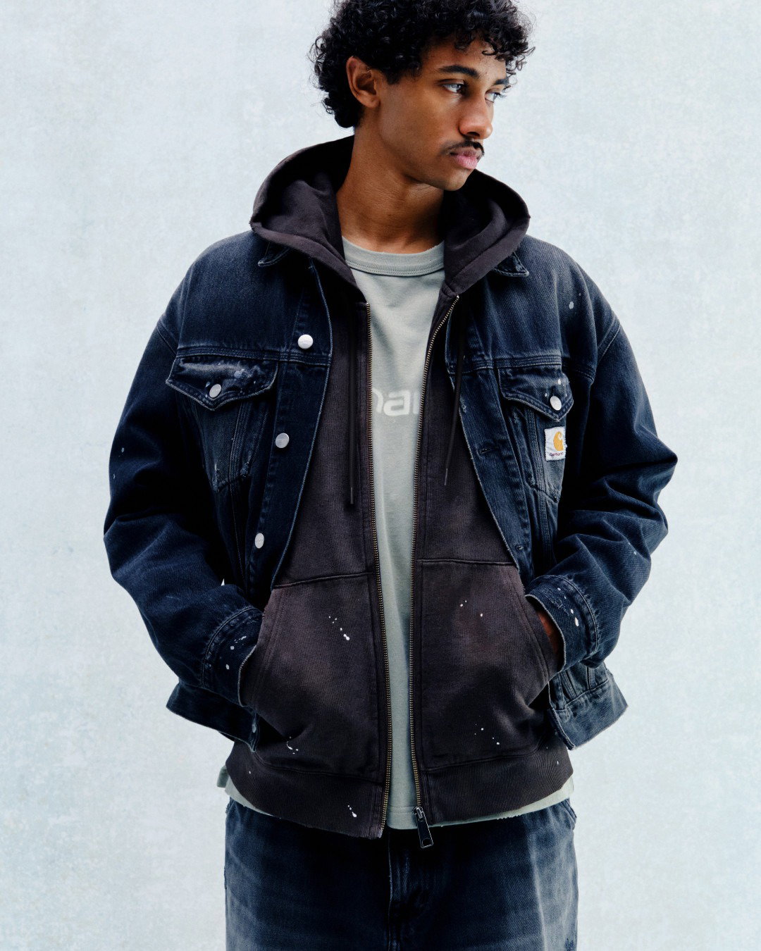 Young man in layered dark denim jacket and paint-spattered brown hoodie, hands in pockets, looking right against pale background