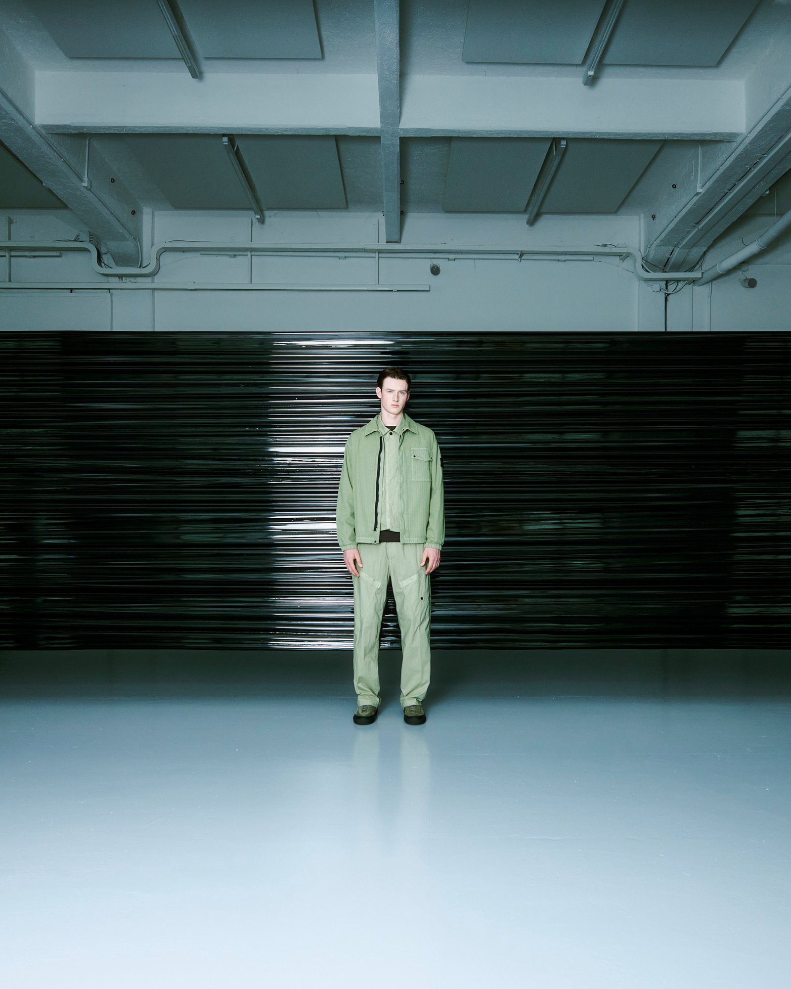 Male model in green jacket and trousers standing centred in an empty industrial space with glossy floor and black shutter