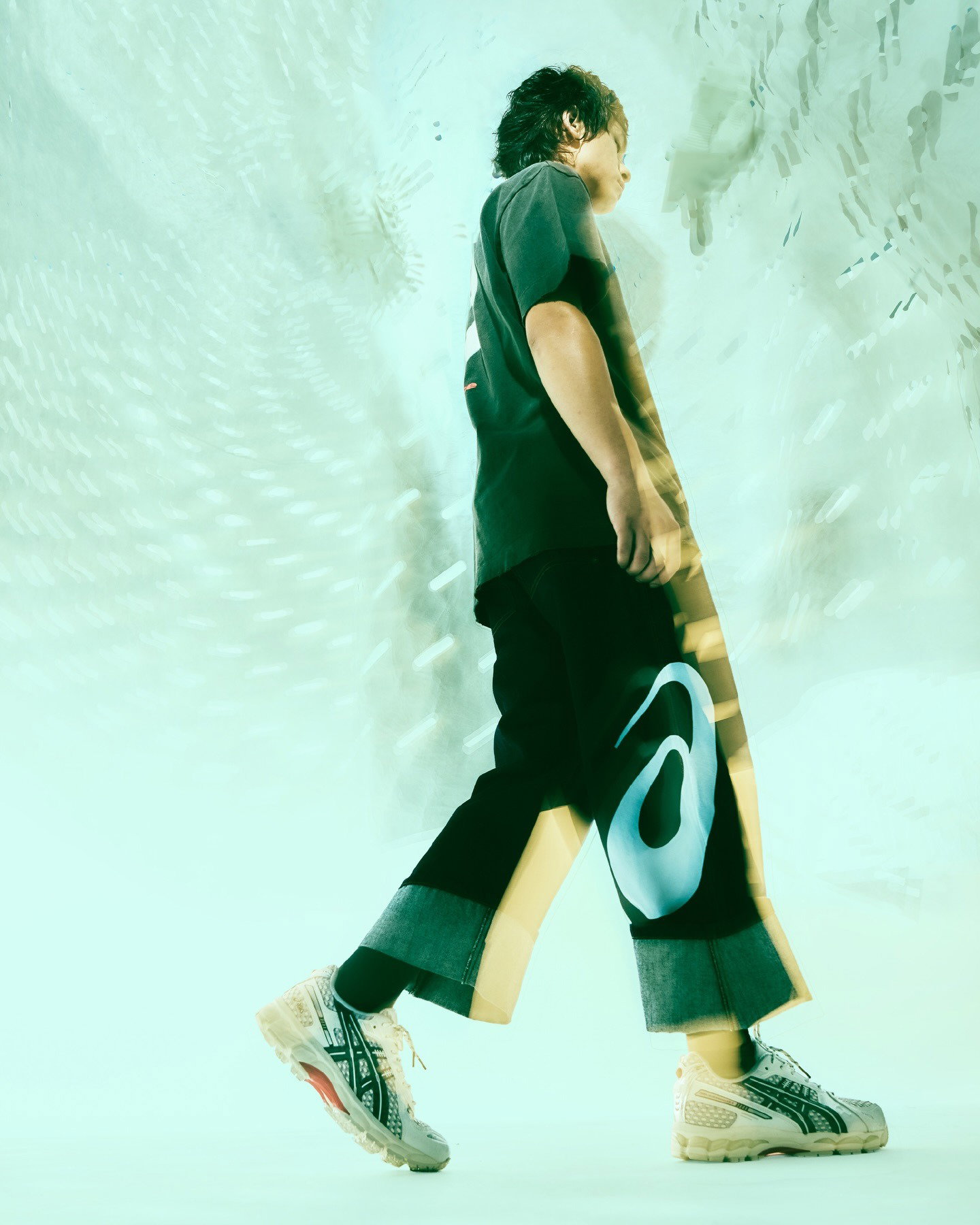 Side view of a young person walking while holding a colourful longboard, wearing cuffed trousers and trainers