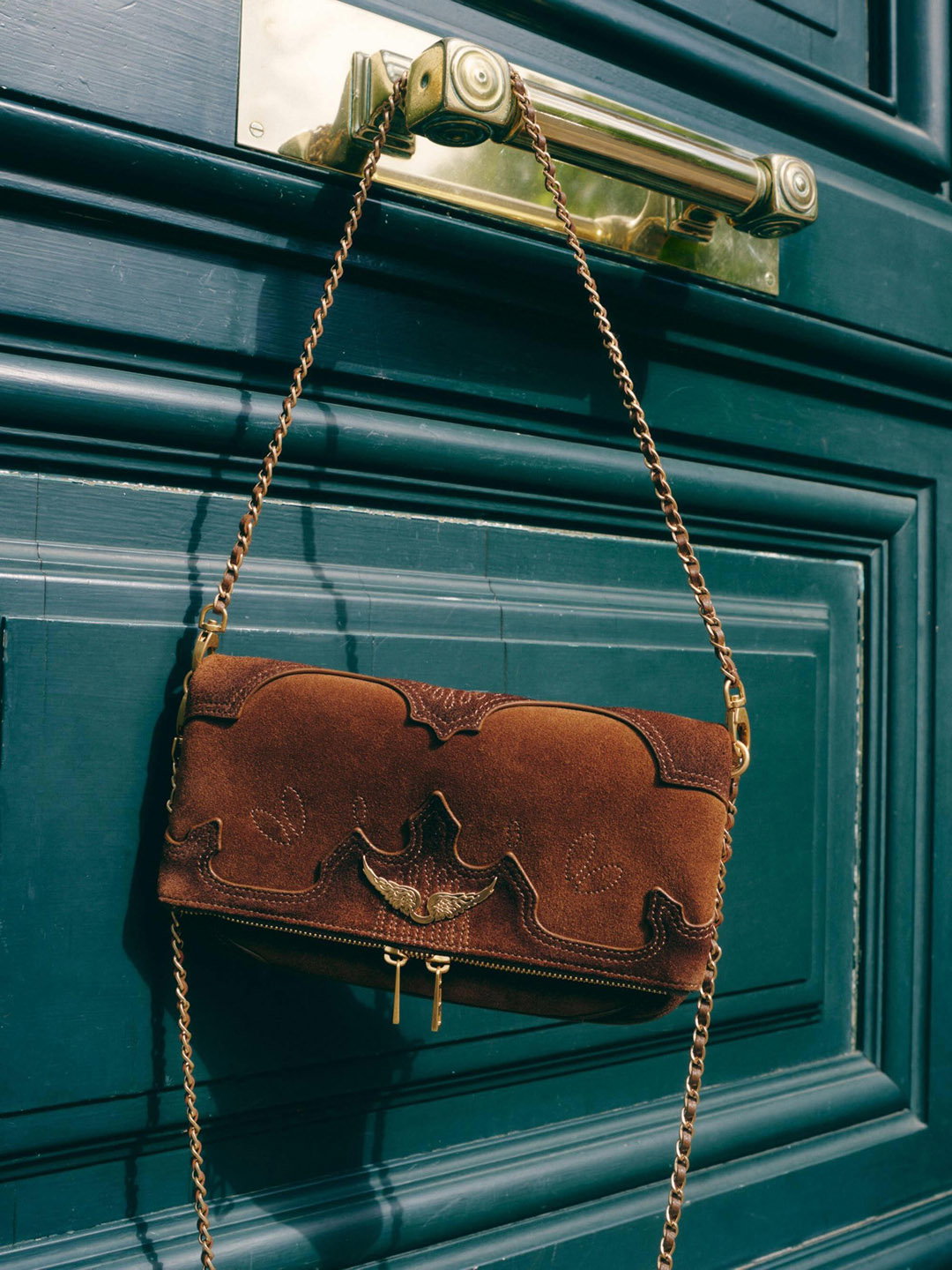 Brown suede chain-strap shoulder bag with decorative stitching and brass clasp hanging from a polished brass door handle on a teal panelled door