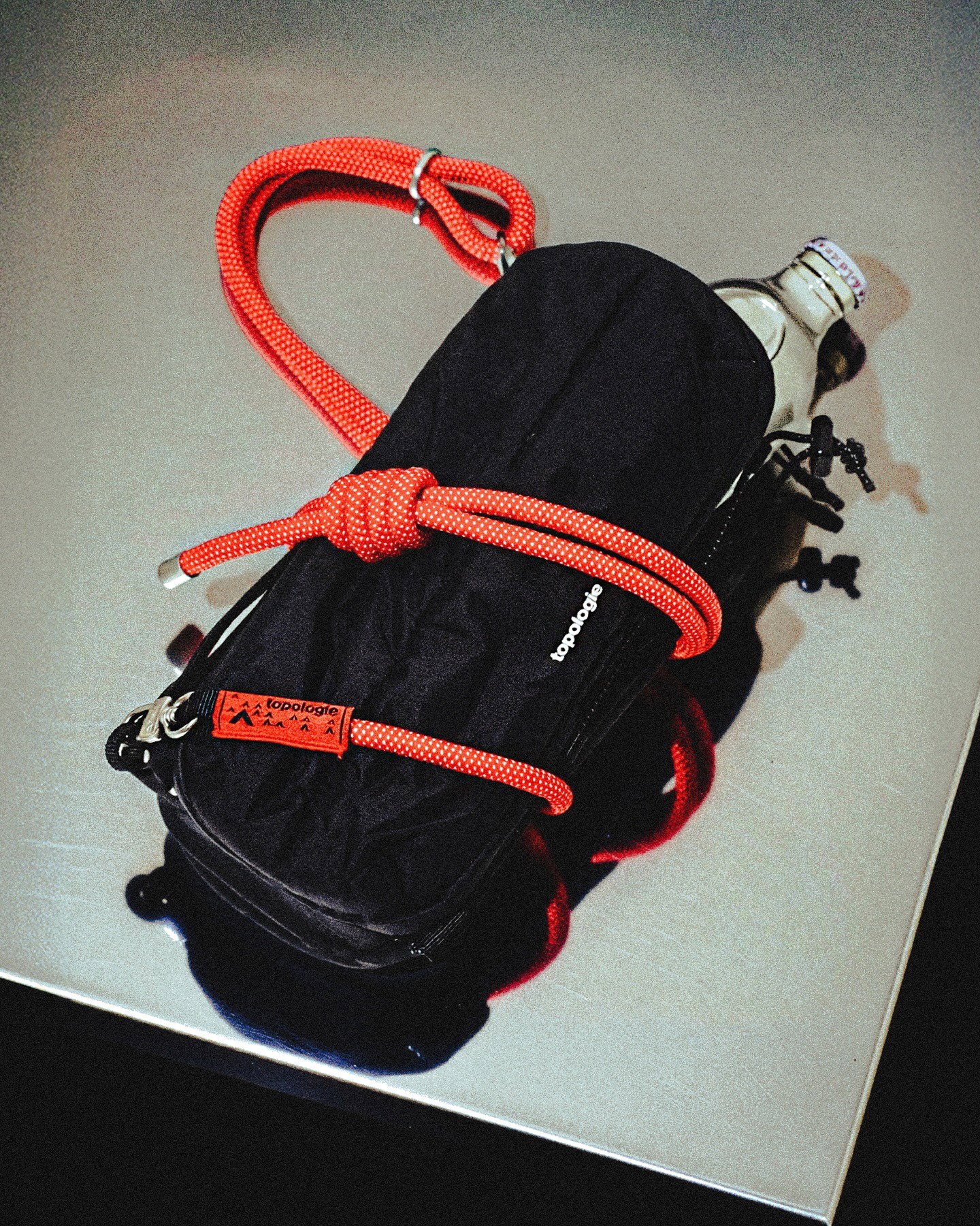 Black Topologie pouch secured with bright red climbing rope strap and a glass water bottle on a reflective tabletop